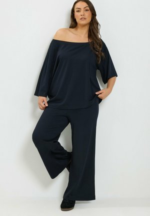 Vrouw draagt losse zwarte off-shoulder top en wijde broekspijpen, staat met één gebogen been tegen een witte muur.