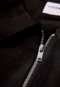 Polo nero in maglia con zip argentata. Presenta un tessuto testurizzato e un'etichetta con la scritta "LINDH" visibile vicino al colletto. La zip è in metallo.
