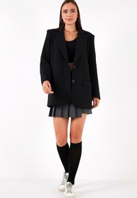 Blazer negru supradimensionat cu închidere cu un singur nasture, asortat cu o fustă gri plisată și șosete negre până la genunchi, și pantofi sport albi.