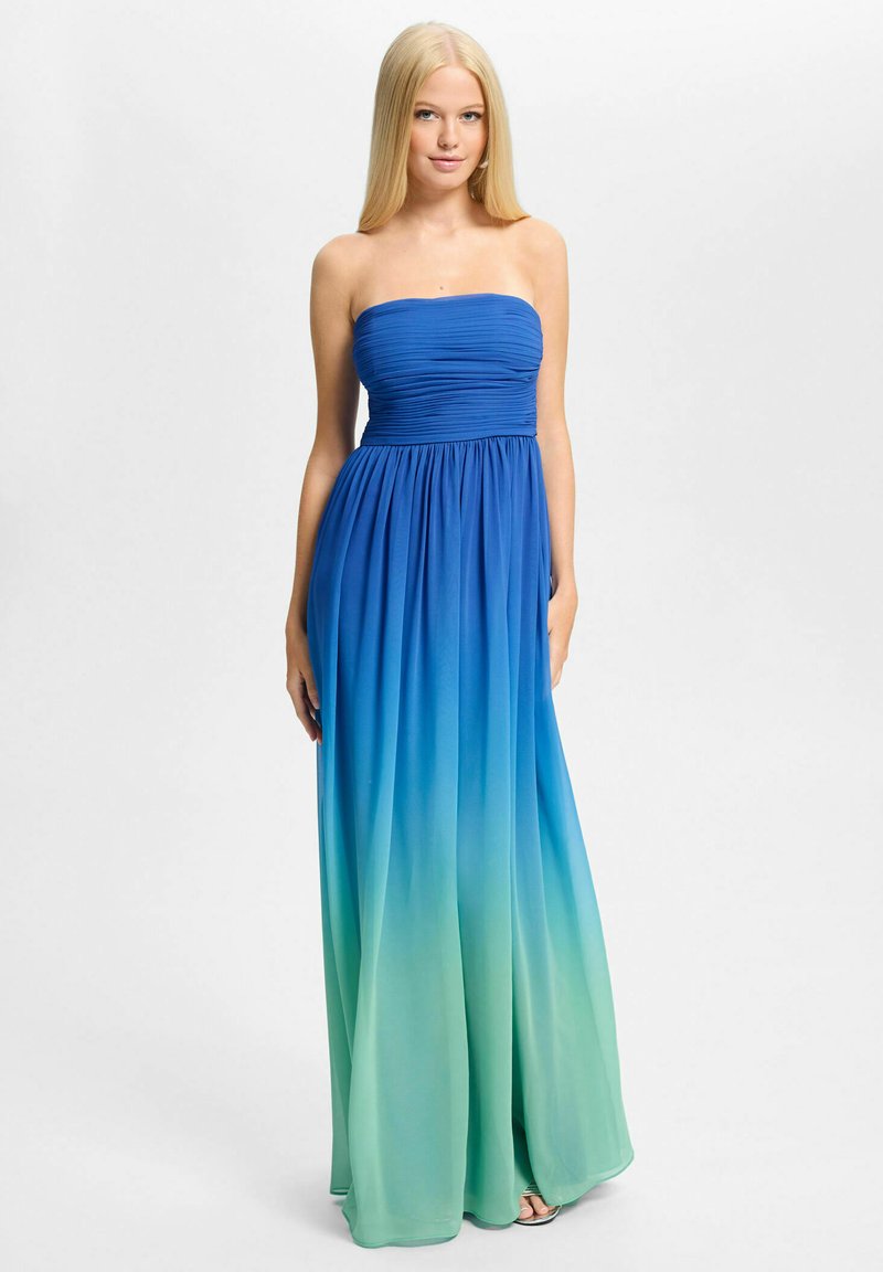 Femme aux longs cheveux blonds portant une robe longue ombrée bleu-vert sans bretelles, debout devant un fond clair uni.
