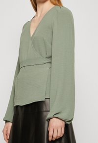 Blouse enveloppante vert clair avec de longues manches bouffantes, finition texturée et détail de taille noué, associée à une jupe en cuir noire.