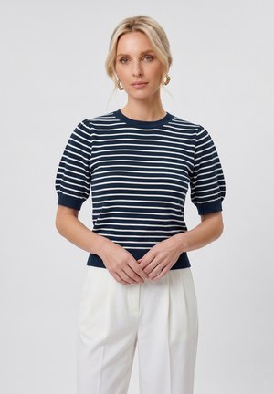 Donna con capelli biondi indossa una maglietta a righe blu navy e bianca a maniche corte e pantaloni bianchi a vita alta, in piedi con le mani intrecciate davanti.