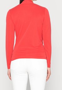Punainen pitkähihainen paita joustavasta kankaasta, jossa on korkea kaulus, istuva siluetti ja sileä tekstuuri. Yhdistetty valkoisiin housuihin.
