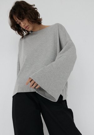 Szary, oversize'owy top z długim rękawem wykonany z miękkiego materiału, o szerokim dekolcie i rękawach w kształcie dzwonu, zestawiony z ciemnymi spodniami.