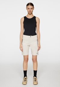 Beiga shorts med sidofickor med dragkedja, i kombination med en svart ärmlös topp. Modellen bär svarta strumpor och bruna skor med färgglada detaljer.