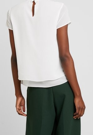 Haut blanc à manches courtes avec un dos en œillet, au design superposé et à la texture légère. Associé à un pantalon large vert, mettant en valeur le contraste de la tenue.