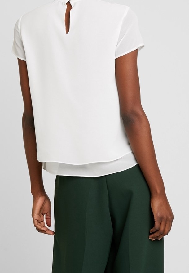 Haut blanc à manches courtes avec un dos en œillet, au design superposé et à la texture légère. Associé à un pantalon large vert, mettant en valeur le contraste de la tenue.