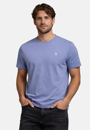 Mann mit kurzen, lockigen braunen Haaren, der ein pastellblaues T-Shirt mit Rundhalsausschnitt und kleinem weißem Logo auf der linken Brust trägt, die Hände in den Taschen einer schwarzen Jeans.