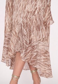 Braun und cremefarbenes gemustertes Kleid mit einem fließenden, asymmetrischen Saum, hergestellt aus leichtem Stoff, kombiniert mit goldenen Knöchelaccessoires.