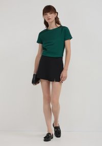 Camicetta verde a maniche corte con collo a giro, abbinata a una minigonna nera plissettata. Mocassini neri e una piccola borsa nera completano il look.