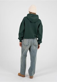 Persoană îmbrăcată cu un hanorac verde închis, blugi albastru deschis, căciulă bej tricotată și cizme maro, stând cu spatele spre înainte pe un fundal alb.