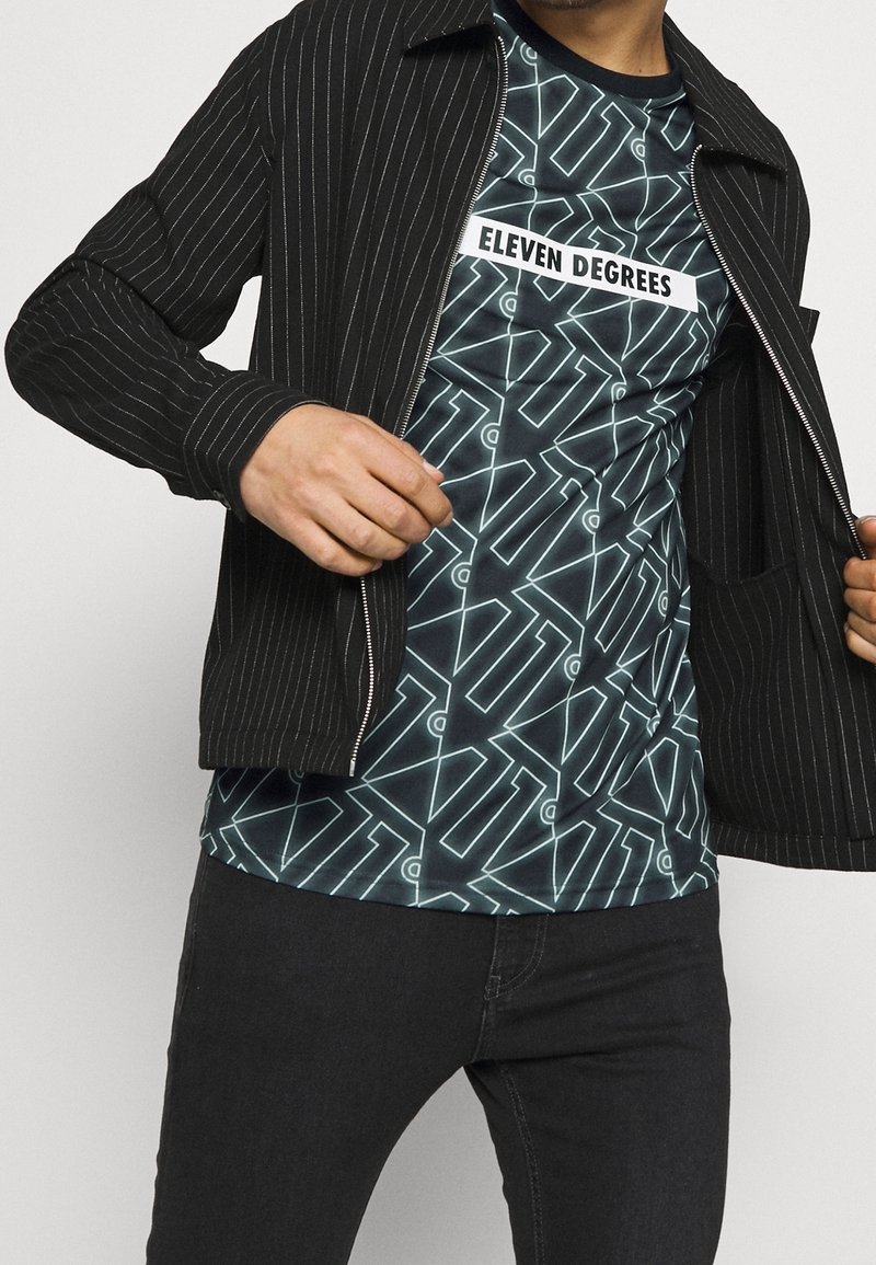 Veste noire à rayures avec fermeture éclair et deux poches sur un T-shirt graphique teal présentant des motifs géométriques et le texte "ELEVEN DEGREES."