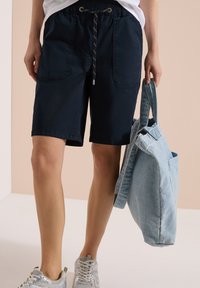 Persona con pantalones cortos negros con cordón y zapatillas blancas, sosteniendo una bolsa tote de mezclilla azul claro a su lado.