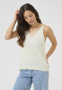 Light beige sleeveless top with a V-neck, made of textured fabric, featuring a subtle pattern and a relaxed fit, paired with light blue jeans.