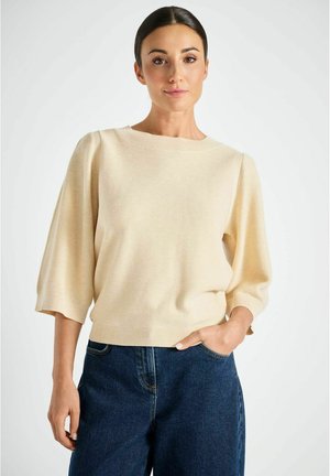 Beige gebreide trui met een ronde hals en driekwart mouwen, gecombineerd met donkerblauwe jeans met wijd uitlopende pijpen. Zachte textuur, casual pasvorm.