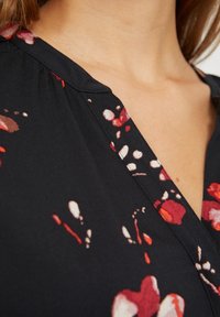 Blusa negra con patrón floral en rojo y blanco. Presenta un escote en V y una tela suave, con pliegues en el hombro para un detalle adicional.