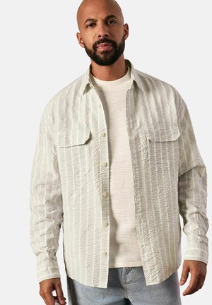 Camisa de botones de rayas ligeramente texturizada en verde claro, llevada abierta sobre un suéter blanco texturizado. Incluye dos bolsillos en el pecho y mangas largas.