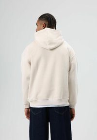 Beige sweatshirt met capuchon en zachte textuur, met verlaagde schouders, geribbelde manchetten en zoom. Draagt over donkerblauwe spijkerbroek, gedeeltelijk zichtbaar.