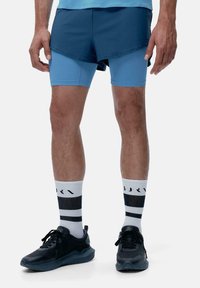 Shorts sportivi blu con un design a strati sopra pantaloni attillati blu chiaro. Sneakers nere con calzini a righe bianche e nere.