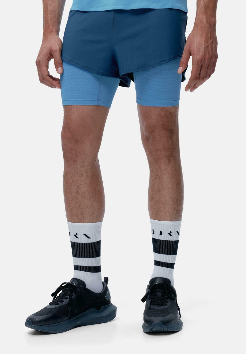 Shorts sportivi blu con un design a strati sopra pantaloni attillati blu chiaro. Sneakers nere con calzini a righe bianche e nere.