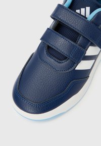 Tmavě modré boty na suchý zip s texturovaným koženým svrškem, síťovaným jazykem a třemi bílými pruhy na boku. Plochá světle modrá podrážka.