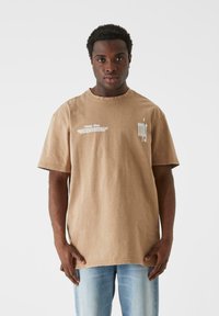 Brown oversized cotton t-shirt with short sleeves, featuring white graphic text on the front. Casual, relaxed fit with a round neckline.