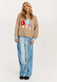 Beżowy sweter dzianinowy z kolorowymi aplikacjami grzybów; niebieskie mankiety. Połączony z jasnoniebieskimi spodniami o szerokich nogawkach i czarnymi butami.
