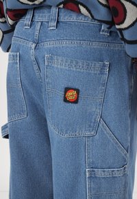 Pantaloni cargo din denim albastru deschis, cu multiple buzunare, o finisare texturată și o etichetă neagră cu logo-ul "Santa Cruz" pe spate.