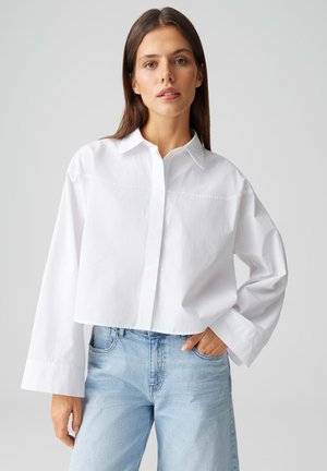 Femme portant une chemise blanche ample à boutons avec des manches larges et un jean bleu clair taille haute, debout avec une main dans la poche.
