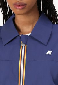 Marineblauwe jas met een kraag, ritssluiting aan de voorkant en strepen in geel, oranje en wit, en een logo op de linkerborst.