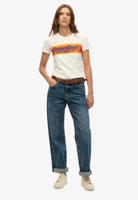 Camiseta de algodón crema con el logo "SuperDry" multicolor, combinada con jeans azules de corte holgado, cinturón marrón y zapatillas crema.