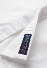 Camisa blanca con cuello abotonado, fabricada en algodón, y una etiqueta azul con texto rojo que detalla la marca y las instrucciones de cuidado.