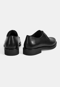 Zwarte leren schoenen met een gladde afwerking, ronde neus en lage, dikke rubberen zolen. Kenmerken zijn minimale veters en geen zichtbare branding.