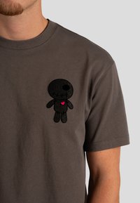 Camiseta de algodón gris con un parche de personaje de dibujos animados negro texturizado y un detalle de corazón rosa en la parte izquierda del pecho. Mangas cortas.