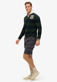 Donkergroene en marineblauwe gestreepte lange mouw polo met een witte kraag, gecombineerd met grijze cargo shorts en beige sneakers.