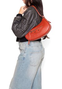Borsa a spalla in pelle rossa con forma curvata, superficie testurizzata e tasca frontale con zip. Abbinata a una giacca bomber nera e jeans azzurri chiari.