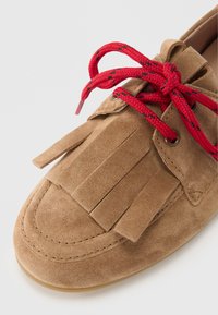 Antilopska mokasina u boji konjaka s crvenim pletenim vezicama. Ima resice na prednjem dijelu i okrugle rupice za vezanje. Dizajn s ravnim potplatom.