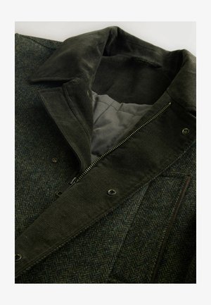 Tummanvihreä villatakki, jossa on kalanruotokuvio, corduroykraavu, vetoketjusta suljettava ja harmaa vuori, jossa on painonapit ja taskut.