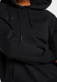 Personne portant un sweat à capuche noir avec cordons de serrage et texte subtilement embossé, bras croisés à la taille, ne montrant que le haut du torse.