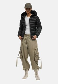 Giacca a vento nera con texture a coste e cappuccio foderato in pelliccia, abbinata a pantaloni cargo beige con tasche oversize e dettagli con coulisse.