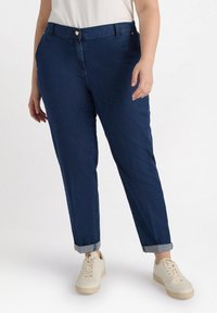 Ciemnoniebieskie dżinsy z podwiniętymi mankietami, pięciopocketowy design i zapięcie na guzik z przodu. Noszone z jasnymi trampkami.