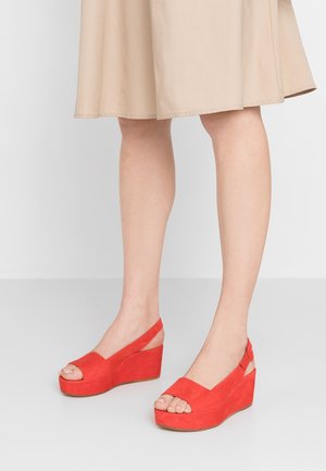 Mujer con falda beige y sandalias de cuña abiertas en rojo brillante, de pie sobre un suelo gris claro.