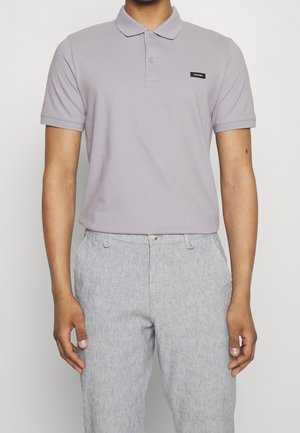 Homme portant un polo gris clair avec le logo Calvin Klein et un pantalon gris clair texturé, debout devant un fond blanc uni.