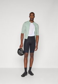 Maillot de vélo vert clair, à manches courtes, associé à un cuissard noir. L'homme tient un casque noir, portant des chaussures et des chaussettes de vélo noires.