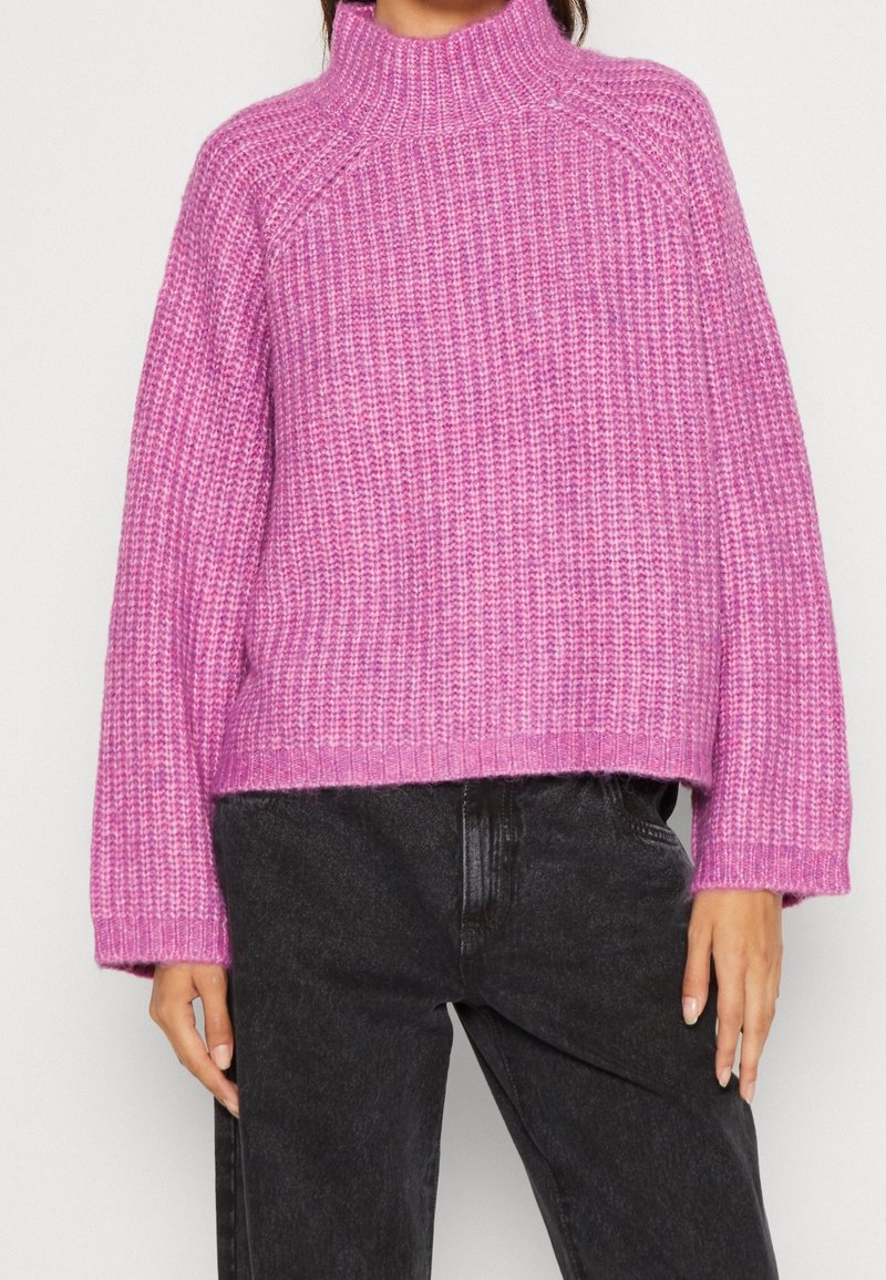 Femme portant un pull rose en grosse maille à col montant et manches longues, associé à un jean en denim noir.