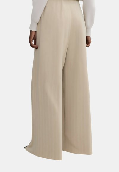 Pantaloni beige a gamba larga indossati da una persona dalla pelle scura, abbinati a una maglia bianca a maniche lunghe, mostrati di spalle.