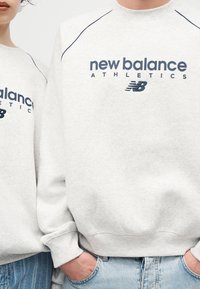 Δύο άτομα που φορούν ανοιχτό γκρι φούτερ New Balance Athletics με μπλε λογότυπο, στέκονται δίπλα δίπλα με τα χέρια στις τσέπες.