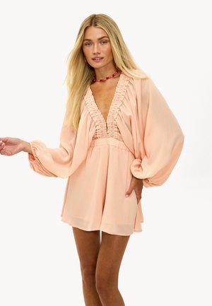 Mujer rubia que lleva un mono suelto de color durazno con mangas largas abullonadas y un profundo escote en V con volantes, accessorized con un collar de cuentas rojas.