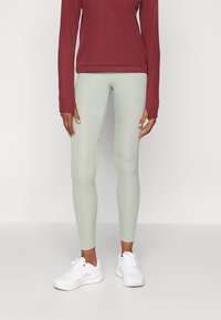 Világoszöld leggings, rugalmas anyagból készült, egy bordó hosszú ujjú felsővel párosítva. Fehér sportcipő egészíti ki az összeállítást.