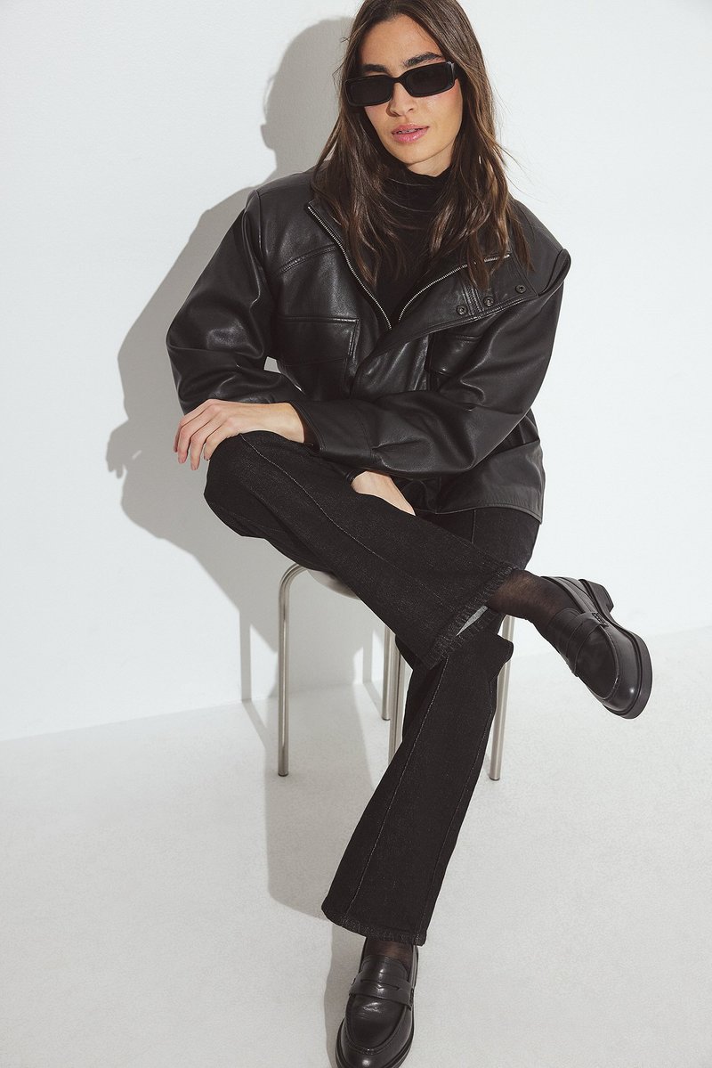 Sort læderjakke med krave, sort turtleneck, mørk denim flared jeans og sorte loafers. Model sidder på en metalstol mod en hvid baggrund.
